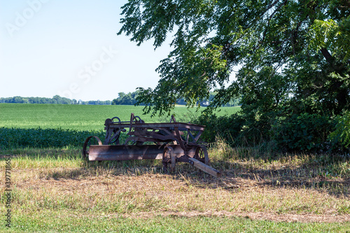 Landscape view of an antique tractor-drawn road grader farm implement, under a tree along the edge of a scenic crop field with blue sky