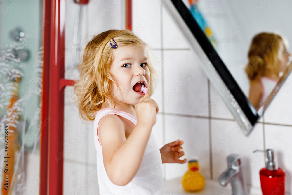 Cute adorable toddler girl holding toothbrush and brushing first teeth in bathroom after sleeping. Gorgeous baby child learning to clean milk tooth. Morning healthy hygiene routine for children Cute adorable toddler girl holding toothbrush and brushing first teeth in bathroom after sleeping. Gorgeous baby child learning to clean milk tooth. Morning healthy hygiene routine for children