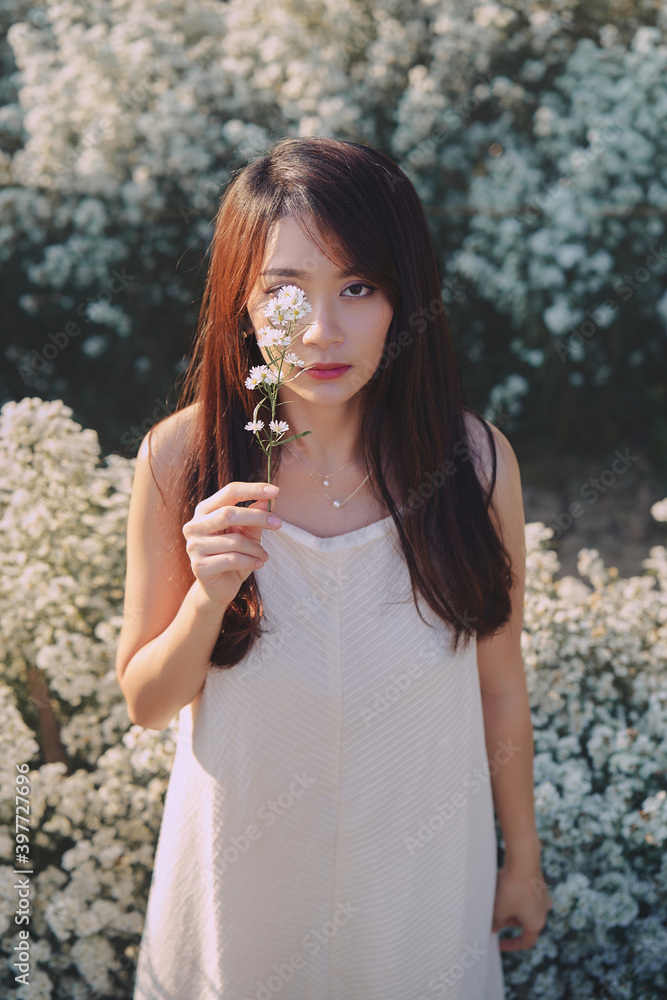 Asian woman in white dress covering her right eye by flower in the garden of white flowers