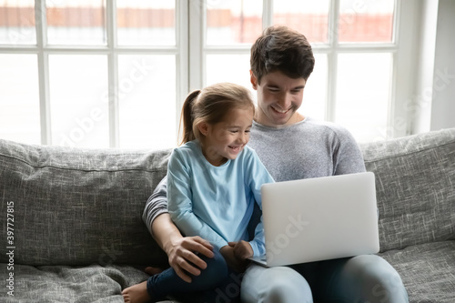 Happy young father cuddling small cute child daughter, teaching using computer application, relaxing together on couch. Smiling little preschool baby girl enjoying watching cartoons online with daddy.