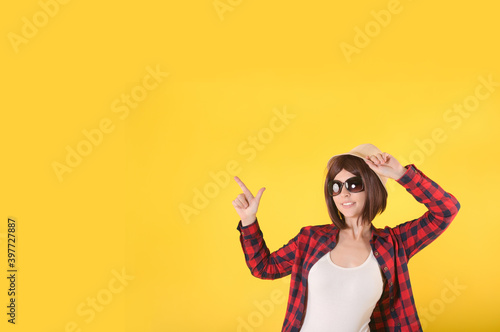 A young girl with short hair in a red checked shirt  straw hat and sunglasses points to an empty space for text on a yellow background and smiles. Half body
