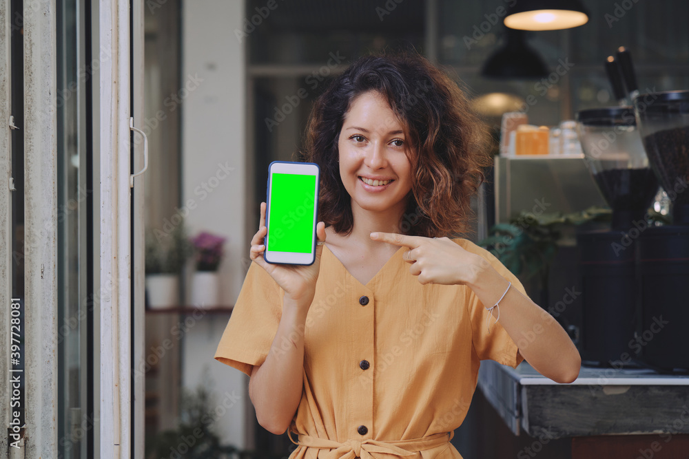 Mockup image of a woman wearing yellow dress holding and showing mobile phone with blank green screen at coffee shop