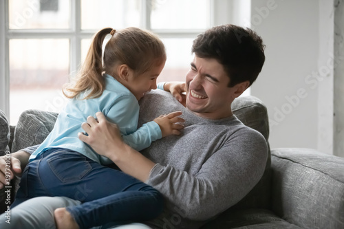 Joyful small preschool baby girl tickling laughing young father, having fun together on sofa at home. Happy millennial daddy playing with overjoyed little daughter, enjoying weekend time indoors.
