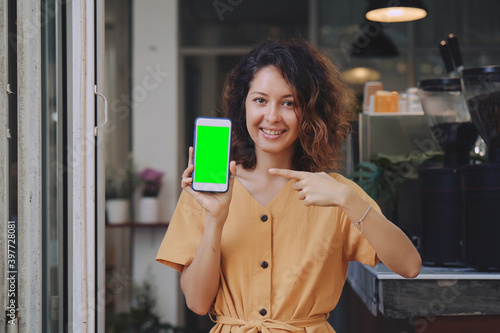 Mockup image of a woman wearing yellow dress holding and showing mobile phone with blank green screen at coffee shop