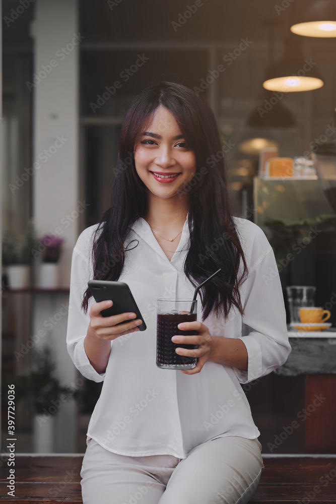 young attractive Asian woman typing a message on her smart-phone in a coffee-shop. She is holding a glass of iced coffee