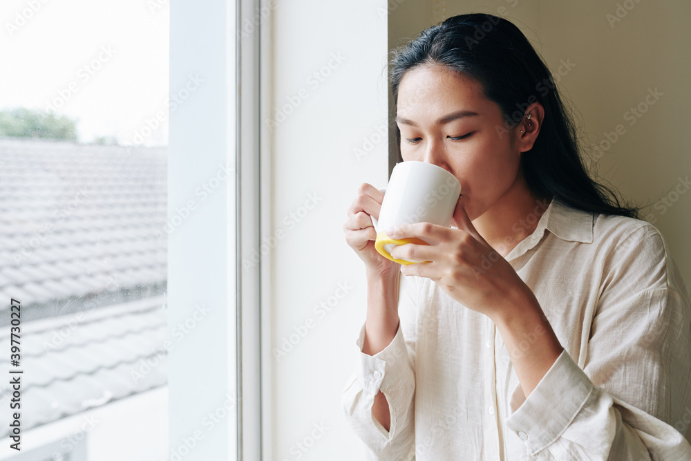 Beautiful young Chiense woman enjoying big mug of morning coffee when standing at window