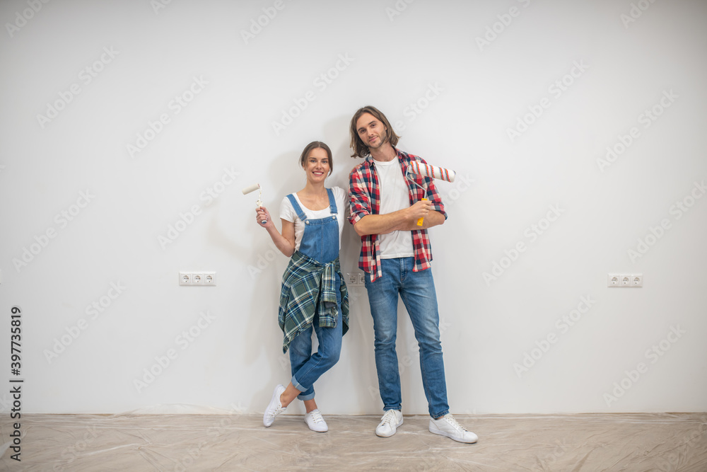 Young couple standing near the wall with paint rollers and hands Young couple standing near the wall with paint rollers and hands