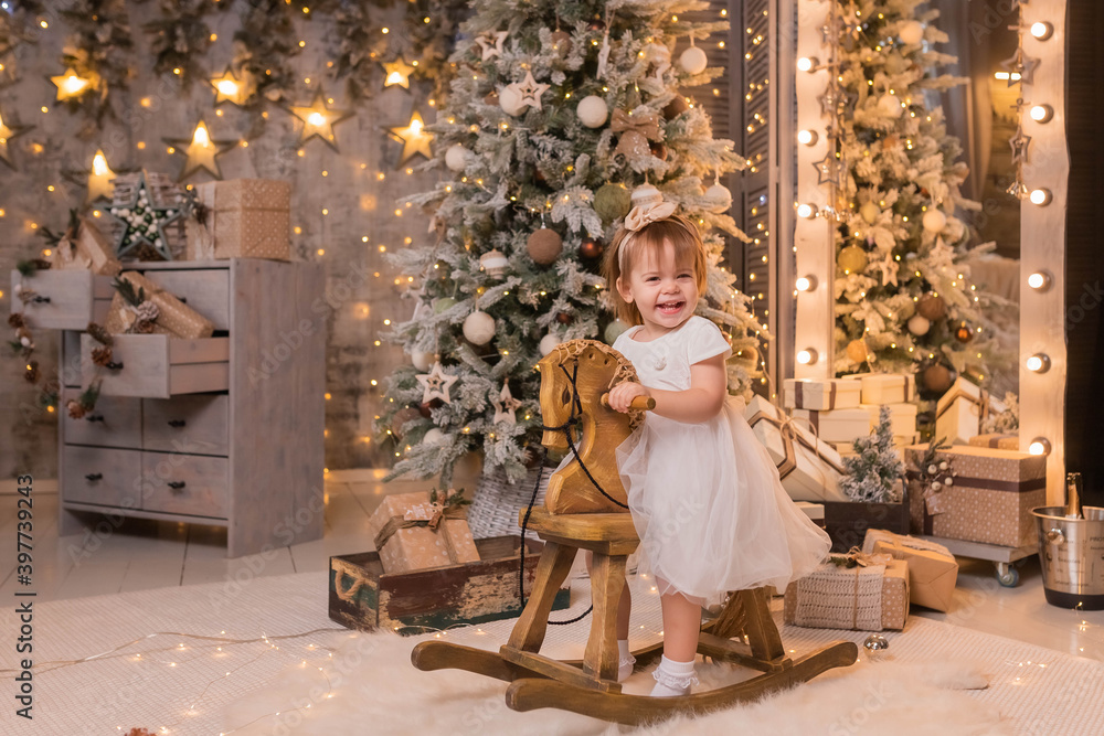 Selective focus. Close-up portrait of a little girl riding a horse on a wooden vintage rocking chair against the background of a Christmas tree. Christmas and New Year. Holiday concept. 