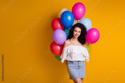 Photo of pretty cute brown curly girl hold many balloons behind back wear top skirt isolated on yellow color background