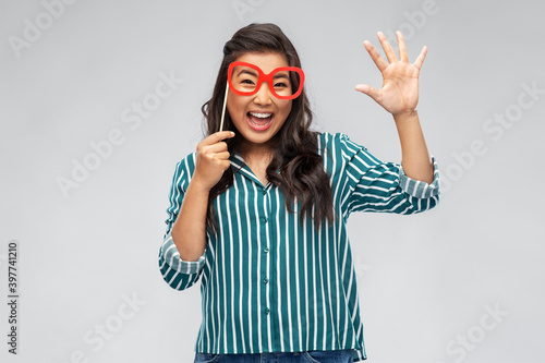 party props, photo booth and people concept - happy asian young woman with big glasses making faces and waving hand over grey background