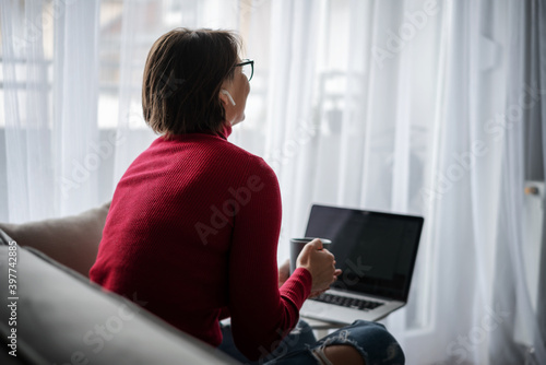 Beautiful smiling happy woman in glasses in a red turtleneck at home sitting at home on a sofa in front of a laptop screen with a mug of coffee