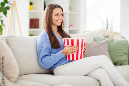 Profile side view portrait of lovely cheerful girl sitting on divan eating corn watching tv in light flat house apartment indoor