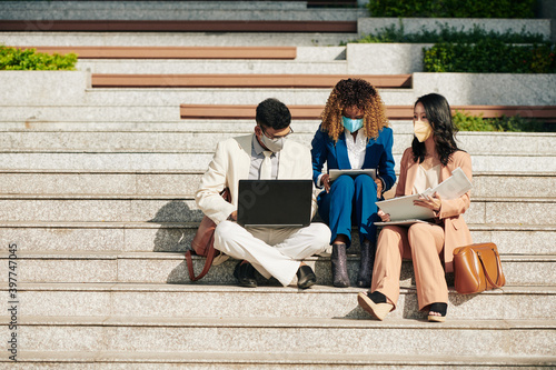 Multi-ethnic business team in medical masks sitting on steps with laptop and documentss and working together on project