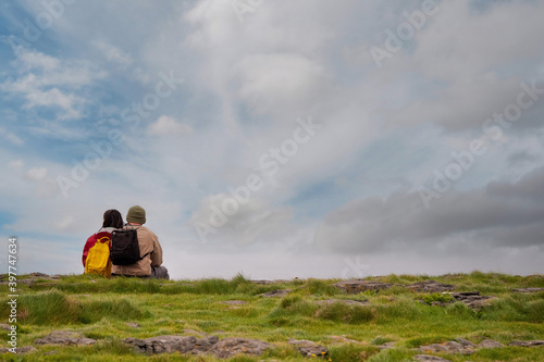 Man and woman tourists with backpacks sitting close to each other on a green grass, looking at a beautiful blue cloudy sky, their back to camera. Concept love and mutual hobby and interests.