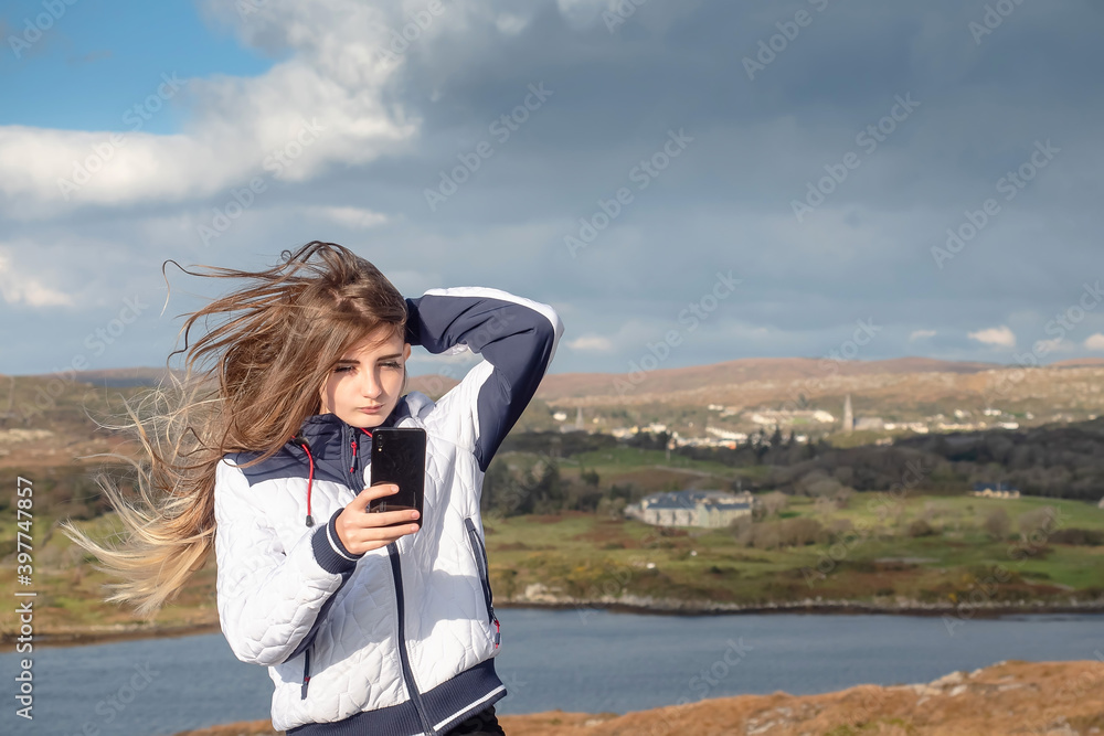Slim teenager girl with long hair taking selfie on her smart phone with beautiful scenery in the background. West coast of Ireland. Clifden area. Model dressed in white jacket. Cloudy sky.