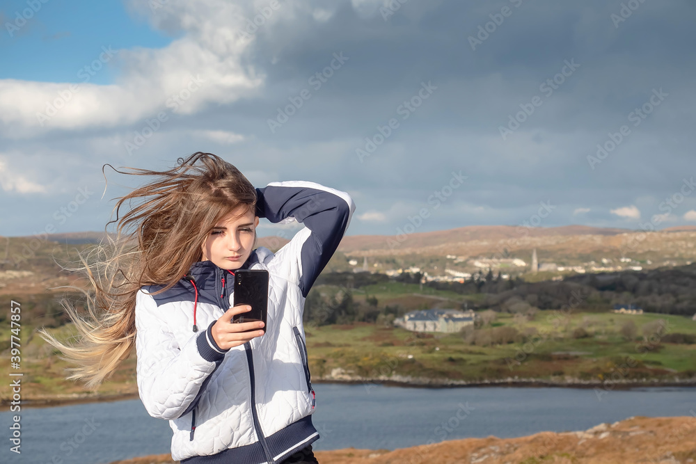 Slim teenager girl with long hair taking selfie on her smart phone with beautiful scenery in the background. West coast of Ireland. Clifden area. Model dressed in white jacket. Cloudy sky. Slim teenager girl with long hair taking selfie on her smart phone with beautiful scenery in the background. West coast of Ireland. Clifden area. Model dressed in white jacket. Cloudy sky.