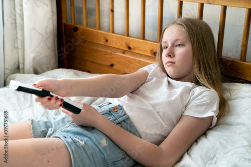 a 13 year old girl is lying on the bed and holding the TV remote in the bedroom near the window
