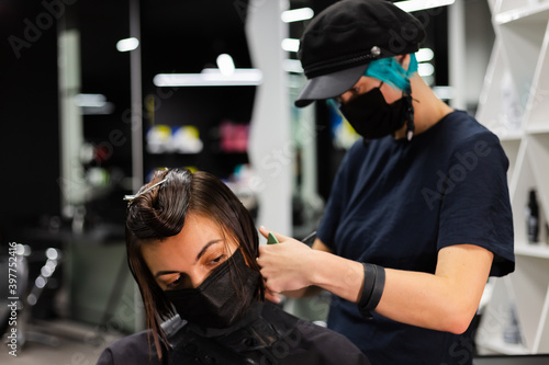 A professional girl hairdresser makes a client haircut. The girl is sitting in a mask in beauty the salon. Social distance. Protective measures. Both woman wearing face masks. 