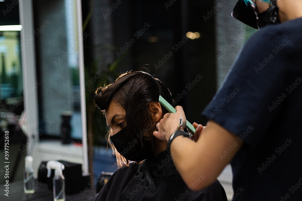 A professional girl hairdresser makes a client haircut. The girl is sitting in a mask in beauty the salon. Social distance. Protective measures. Both woman wearing face masks.
  