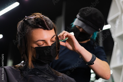 A professional girl hairdresser makes a client haircut. The girl is sitting in a mask in beauty the salon. Social distance. Protective measures. Both woman wearing face masks.    © Анастасия Каргаполов