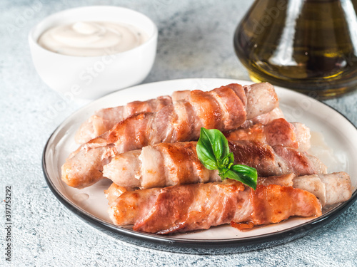 Ready-to-eat pigs sausages wrapped in bacon on plate. Fried savory sausages wrapped in bacon served fresh green basil leaves with sauce on background. Shallow DOF. Copy space for text.