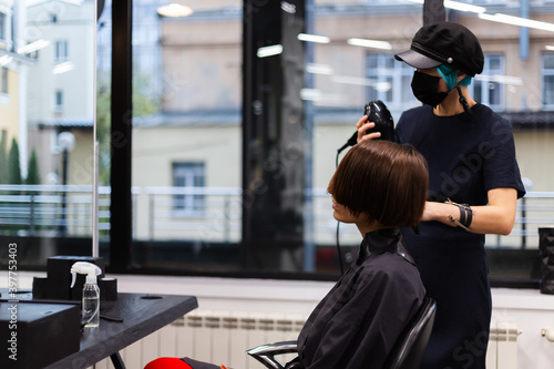 A professional girl hairdresser in face mask makes a client trendy stylish short hair cut. The girl is sitting in stylish beauty salon. Social distance. Protective measures. 
