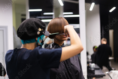A professional girl hairdresser in face mask makes a client trendy stylish short hair cut. The girl is sitting in stylish beauty salon. Social distance. Protective measures. 