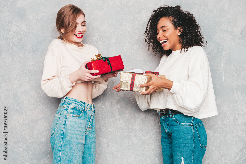 Two young beautiful smiling international hipster female in trendy clothes.Sexy carefree women posing near gray wall.Positive models hugging and giving each other gift boxes.Christmas, x-mas, concept Two young beautiful smiling international hipster female in trendy clothes.Sexy carefree women posing near gray wall.Positive models hugging and giving each other gift boxes.Christmas, x-mas, concept