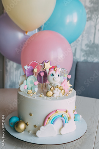 Birthday cake to a girl with a unicorn, vertical frame. Cake for a girl of gentle pastel tones, decorated with unicorn figures with a bear and rainbow, lollipops, on the background - colorful balls
