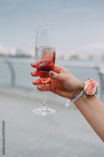 A glass of champagne in a woman's hand,  close-up. The woman's hand, decorated with a wedding bracelet of flowers on a white ribbon, holds a glass of pink champagne