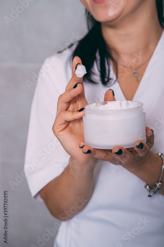 Hands of beautician in cream and with a jar of cream in hand. Beautician holds a jar of facial skin care cream, one finger in white cream, vertical frame close-up