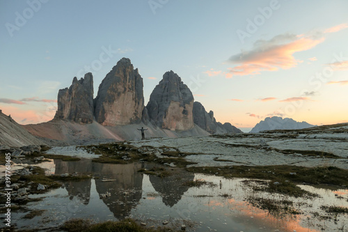 A man enjoying the sunset over the Tre Cime di Lavaredo (Drei Zinnen) mountains in Italian Dolomites. The peaks reflect in a paddle. The mountains are surrounded with orange and pink clouds. Freedom