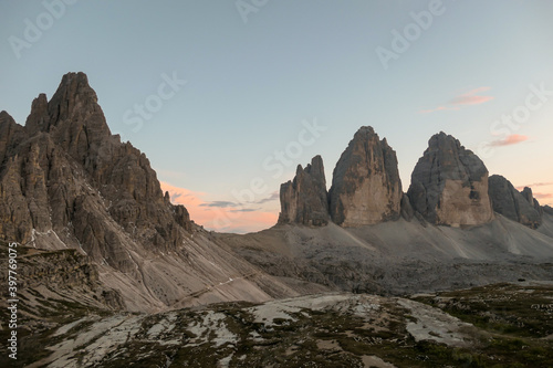 A panoramic capture of the sunset above Tre Cime di Lavaredo (Drei Zinnen) and surrounding mountains in Italian Dolomites. The mountains are surrounded by pink and orange clouds. Golden hour. Serenity