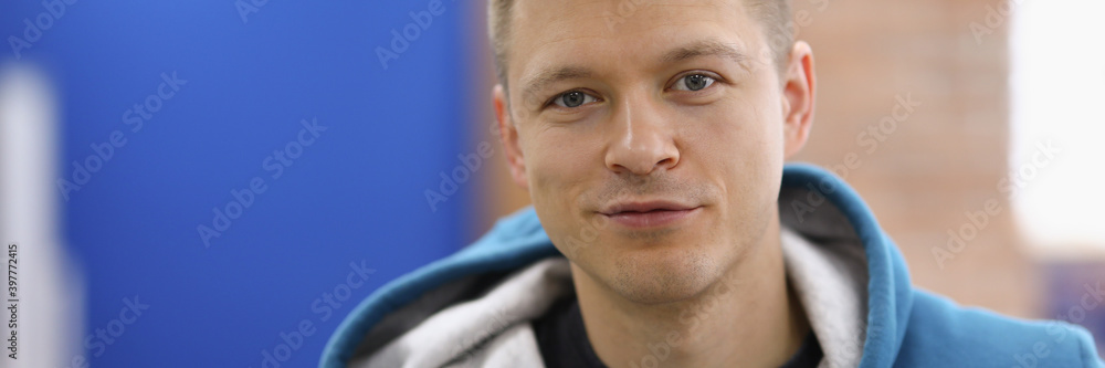 Obraz premium Portrait of smiling cheerful young man posing on camera in room. Happy adult wearing blue hoodie. Copy space in left side. Modeling and happiness concept