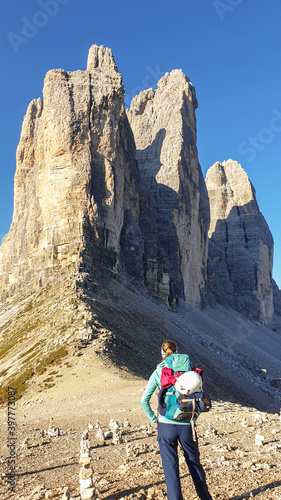 A woman with big hiking backpack enjoying the close up view on the Tre Cime di Lavaredo (Drei Zinnen), mountains in Italian Dolomites. Sharp and high mountain wall. Desolated and raw landscape.