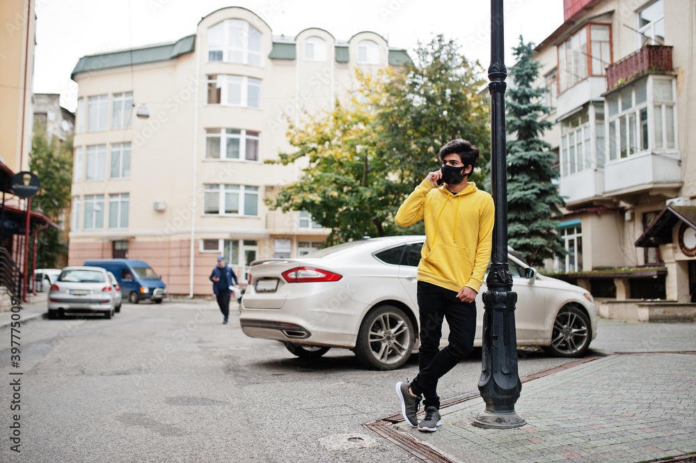 Urban young hipster indian man in a fashionable yellow sweatshirt with mobile phone. Cool south asian guy wear hoodie and black face protect mask, during new normal.