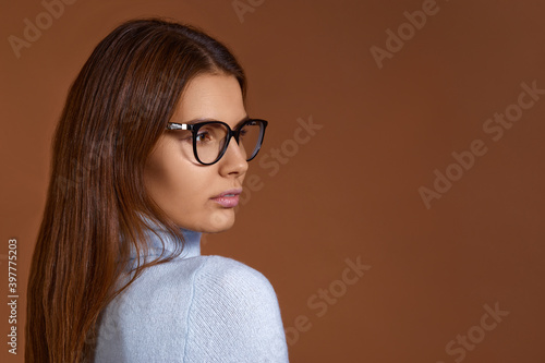 Portrait of a young smart looking european woman with dark long hair wearing eyeglasses and light blue sweater, looks aside, isolated on brown background, copy space on the right.