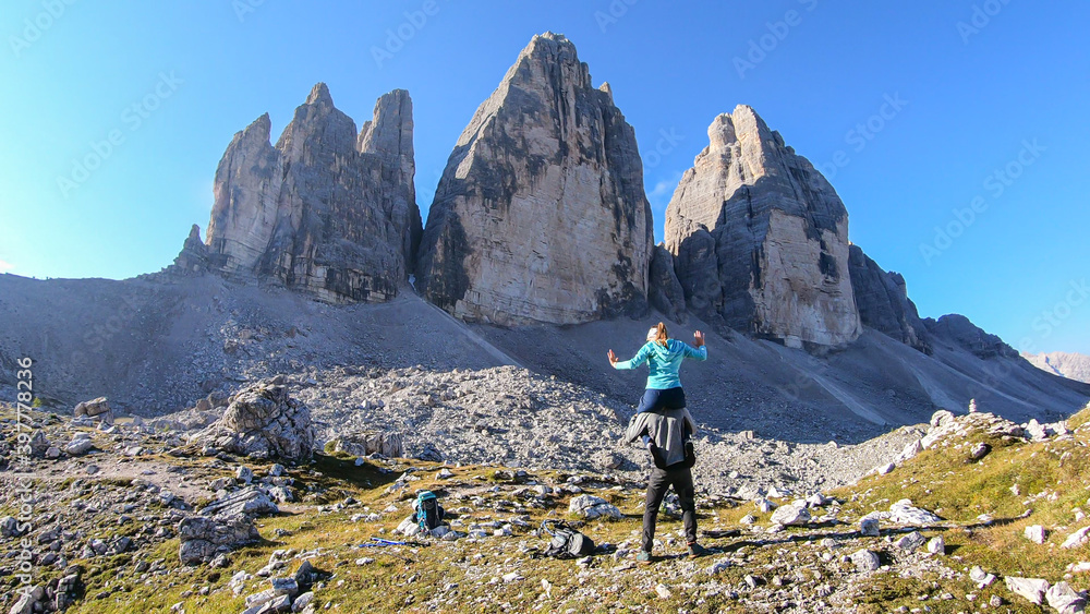 Man carrying woman piggyback with the close up view on the Tre Cime di Lavaredo (Drei Zinnen) in Italian Dolomites. Sharp and high mountain wall. Desolated and raw landscape. Fun and careless moments