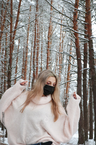 Woman with blond long hair posing in a snowy park. She is wearing an oversized sweater and a protective mask. Deserted forest. Winter landscapes. © Olha