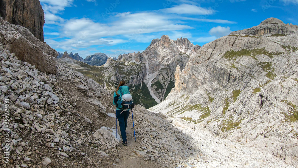 A woman with backpack and sticks hiking on a narrow path in Italian Dolomites. There are sharp and steep mountains around her. Lots of lose stones. Raw and desolated landscape. Following the pathway