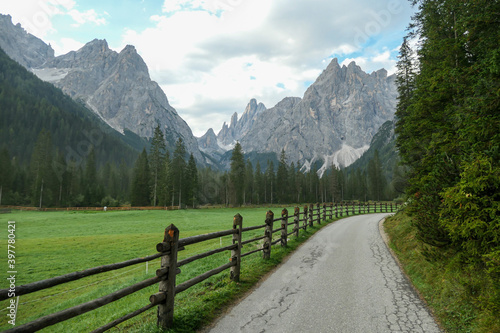 A gravelled road leading along a lush green meadow, secured with a wooden fence, to Italian Dolomites. Morning fog in the valley. Thick forest on the other side of the road. Idyllic landscape