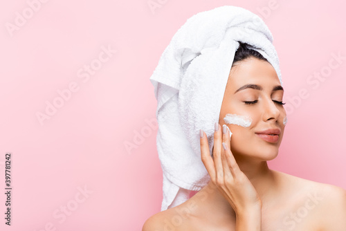 beautiful woman with towel on hair, closed eyes and cosmetic cream on face isolated on pink