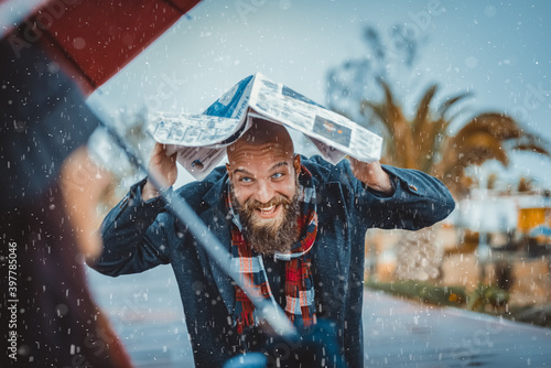 couple of bearded bald man holding a newspaper running under a snow winter storm and caucasian brunette woman wearing warm clothes holding an umbrella. hipster boy and a girl having fun on christmas.