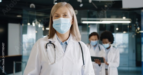 Close up portrait of Caucasian senior female doctor in medical mask standing in hospital looking at camera at work. Healthcare professionals using tablet on background. Coronavirus concept