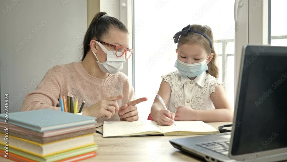 Mom or teacher or tutor helps an elementary school student with her lessons. Both have medical masks. Training during the COVID-19 pandemic. Mom or teacher or tutor helps an elementary school student with her lessons. Both have medical masks. Training during the COVID-19 pandemic.