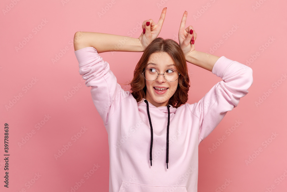 Portrait of funny young woman in eyeglasses and hoodie gesturing horns over head and smiling, looking to side with comical humorous expression. Indoor studio shot, isolated on pink background Portrait of funny young woman in eyeglasses and hoodie gesturing horns over head and smiling, looking to side with comical humorous expression. Indoor studio shot, isolated on pink background