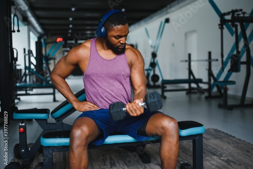 Young African American man sitting and lifting a dumbbell close to the rack at gym. Male weight training person doing a biceps curl in fitness center