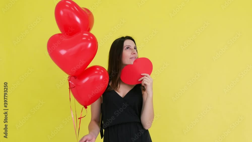 Portrait of a happy woman with a red heart, holding in her hands, covering her face with a red paper heart, smiling at the camera on a yellow background. Emotions Positive girl Flirting. Valentine's