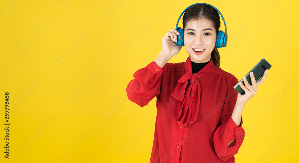 Asian women in the happy mood hold the phone and put the wireless headphone with red dress. listening music from Mobile or studying E-leanning from mobile over yellow background with copy space.
