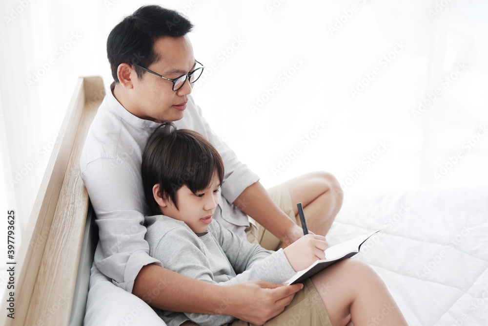 Smiling Asian father and son child is writing a book and doing homework together on bed in bedroom. Family and education on holiday concept. Smiling Asian father and son child is writing a book and doing homework together on bed in bedroom. Family and education on holiday concept.